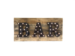 A rustic wooden sign featuring the word 'BAR' in large, black marquee-style letters with warm white light bulbs, mounted on a weathered brown wood plank background.